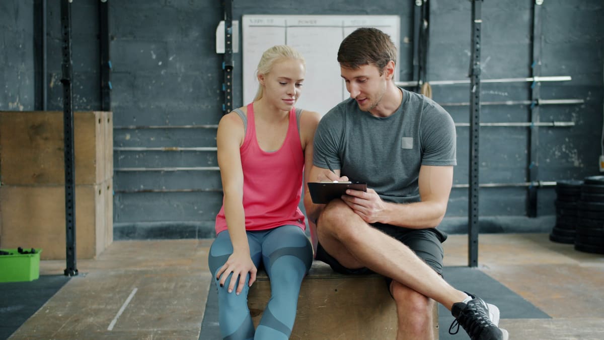 Personal trainer e aluna sentados lado a lado, olhando juntos uma ficha na prancheta