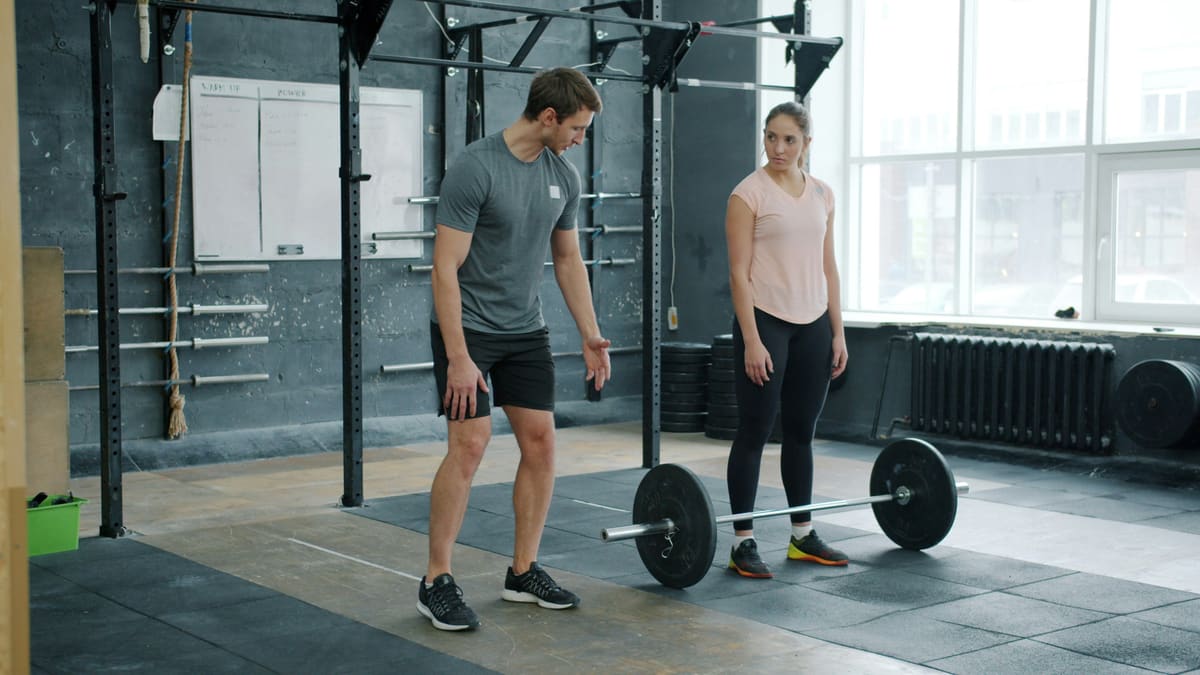Personal trainer e aluna conversando em pé ao lado de uma barra apoiada no chão
