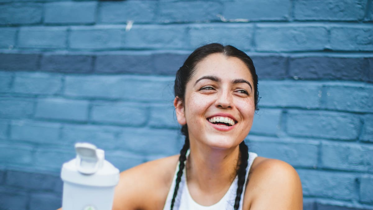 Mulher sorrindo diante de uma parede de tijolos azuis, com uma coqueteleira ao lado